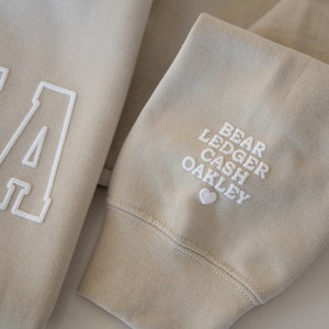May include: A beige sweatshirt with a large white letter "A" on the front and the names "Bear Ledger Cash Oakley" embroidered on the sleeve with a small heart.