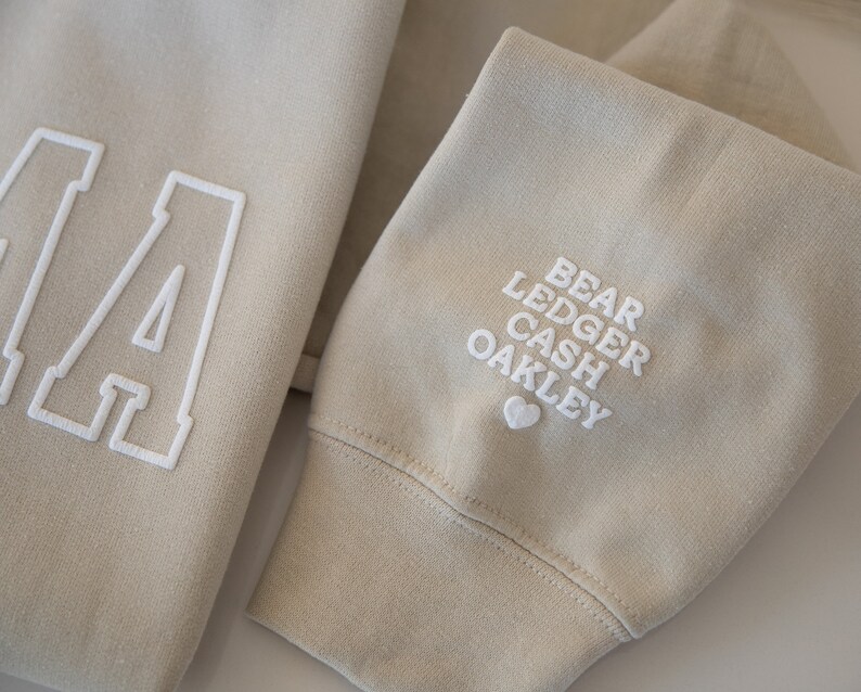 May include: A beige sweatshirt with a large white letter "A" on the front and the names "Bear Ledger Cash Oakley" embroidered on the sleeve with a small heart.