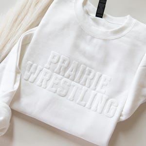 May include: A white long-sleeve crewneck sweatshirt featuring the text PRAIRIE WRESTLING embossed in a raised, tonal font across the chest. The garment has a ribbed collar and cuffs, with a clean, monochromatic design throughout the fabric.