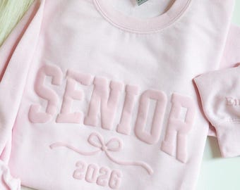 Op de afbeelding: Een lichtroze sweatshirt met het woord "SENIOR" in verhoogde, bijpassende roze letters. Daaronder een strik en het jaar "2026". De sweatshirt heeft een ronde hals en lange mouwen.