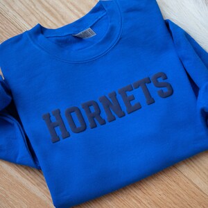 Puede incluir: Una sudadera azul real con la palabra "HORNETS" impresa en azul oscuro en la parte delantera.