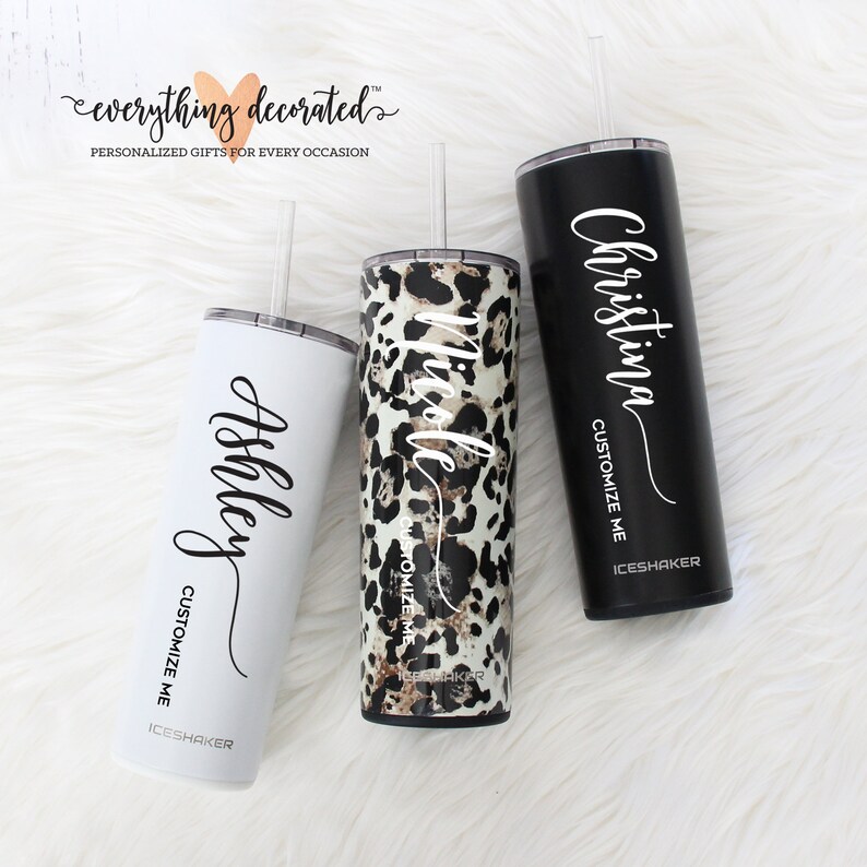 May include: Three stainless steel tumblers with personalized names, "Ashley", "Nicole", and "Christina". Each tumbler has a black lid and a straw. The tumbler on the left is white, the middle tumbler has a leopard print design, and the tumbler on the right is black.