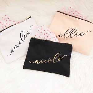 Puede incluir: Tres bolsas de maquillaje personalizadas con letras doradas. Las bolsas son blancas, negras y melocot&oacute;n. Los nombres en las bolsas son "emelia", "ellie" y "nicole".