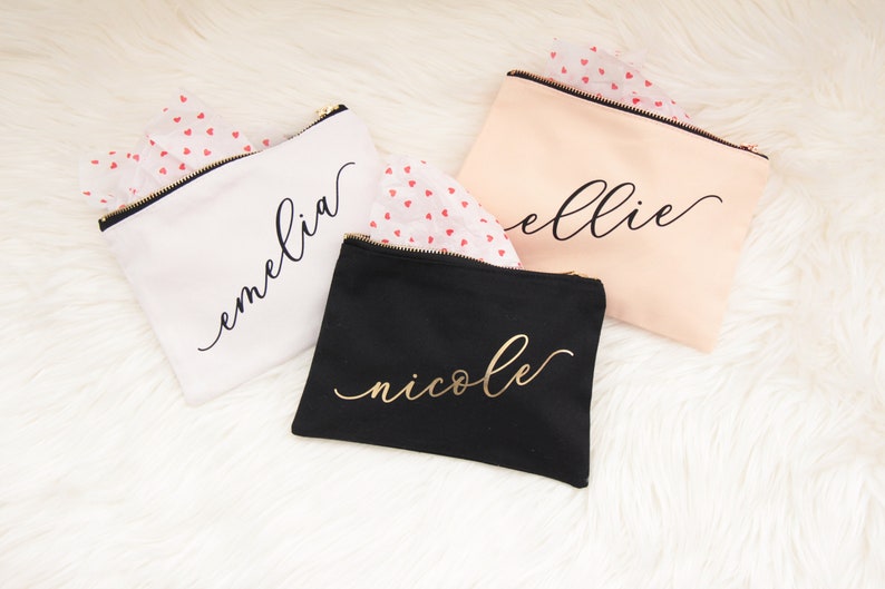 Puede incluir: Tres bolsas de maquillaje personalizadas con letras doradas. Las bolsas son blancas, negras y melocot&oacute;n. Los nombres en las bolsas son "emelia", "ellie" y "nicole".