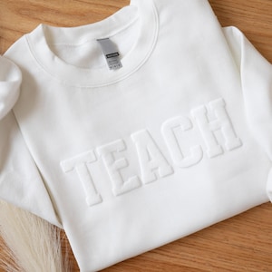 Puede incluir: Una sudadera blanca de cuello redondo con la palabra "TEACH" en relieve en el pecho. La sudadera est&aacute; hecha de un material suave y se muestra sobre una superficie de madera. Las mangas est&aacute;n dobladas y la est&eacute;tica general es limpia y sencilla.