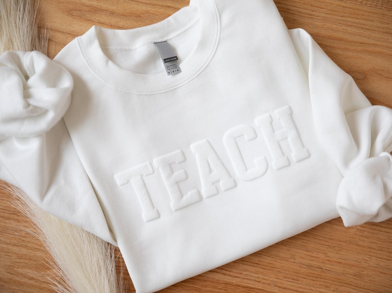 Puede incluir: Una sudadera blanca de cuello redondo con la palabra "TEACH" en relieve en el pecho. La sudadera est&aacute; hecha de un material suave y se muestra sobre una superficie de madera. Las mangas est&aacute;n dobladas y la est&eacute;tica general es limpia y sencilla.