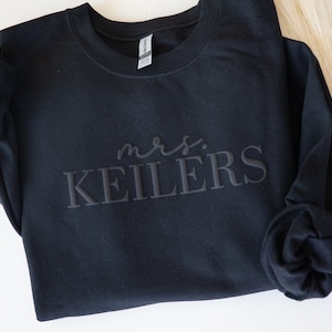K&ouml;nnte beinhalten: Schwarzes Sweatshirt mit dem Text "mrs. KEILERS" in einem Ton-in-Ton-Design. Das lang&auml;rmlige Sweatshirt ist aus einem weichen Material gefertigt. Das Gildan-Etikett ist innen am Kragen sichtbar.