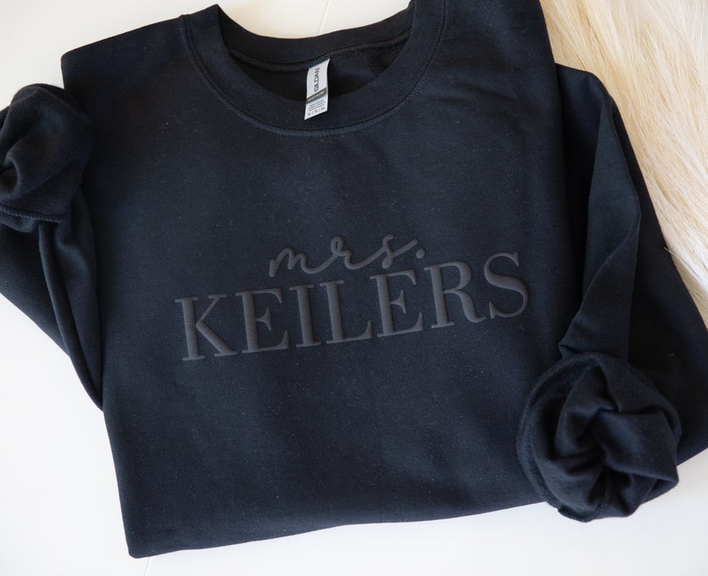 K&ouml;nnte beinhalten: Schwarzes Sweatshirt mit dem Text "mrs. KEILERS" in einem Ton-in-Ton-Design. Das lang&auml;rmlige Sweatshirt ist aus einem weichen Material gefertigt. Das Gildan-Etikett ist innen am Kragen sichtbar.