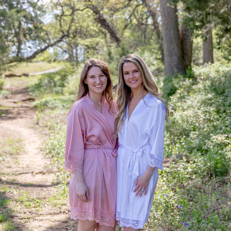 May include: Two women wearing matching robes with lace trim. One robe is pink and the other is white. They are standing in a wooded area with a dirt path behind them.