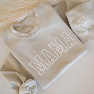 May include: A light beige sweatshirt with the word "MAMA" printed in white on the front. The sweatshirt has a crew neck and long sleeves.