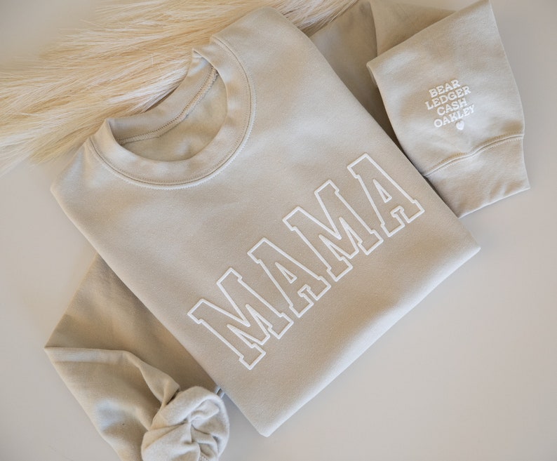 May include: A light beige sweatshirt with the word "MAMA" printed in white on the front. The sweatshirt has a crew neck and long sleeves.