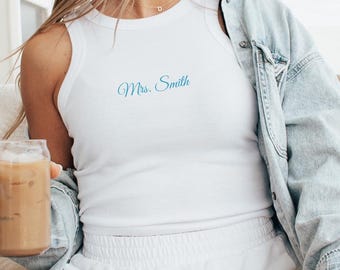May include: White tank top with the text "Mrs. Smith" in a teal script. The top is sleeveless and has a scoop neck. The person is wearing a light blue denim jacket and white shorts.