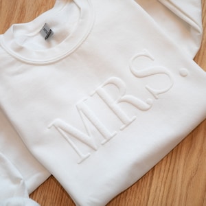 K&ouml;nnte beinhalten: Wei&szlig;es Sweatshirt mit Rundhalsausschnitt und dem gepr&auml;gten Wort &bdquo;MRS.&ldquo; in einem strukturierten, erhabenen Design. Das Sweatshirt ist auf einer Holzoberfl&auml;che gefaltet. Das Gildan-Etikett ist auf der Innenseite des Kragens sichtbar.