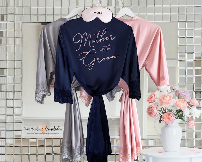 May include: Three satin robes with lace trim, one navy blue with the text "Mother of the Groom" in rose gold lettering, one gray, and one pink. The robes are hanging on hangers with a pink eye mask that says "MOM" on it.