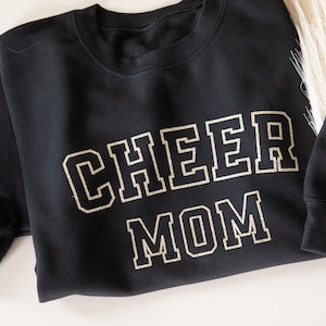 May include: Black sweatshirt with gold glitter lettering that reads "CHEER MOM". The sweatshirt has a small gold glitter bow on the sleeve with the word "HADLEY" above it.