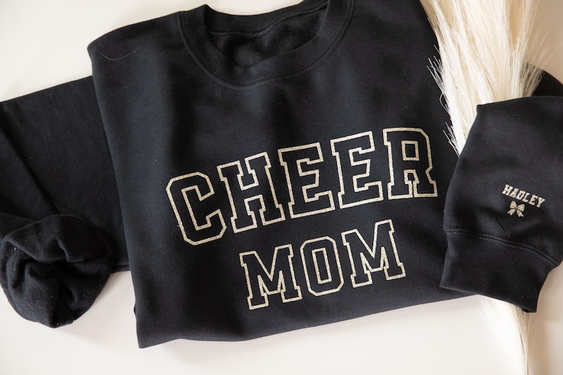 May include: Black sweatshirt with gold glitter lettering that reads "CHEER MOM". The sweatshirt has a small gold glitter bow on the sleeve with the word "HADLEY" above it.