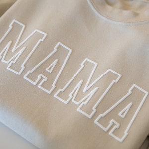 May include: A beige sweatshirt with the word "MAMA" in white outlined letters.