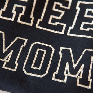 May include: A black shirt with the words "Cheer Mom" in silver glitter lettering.