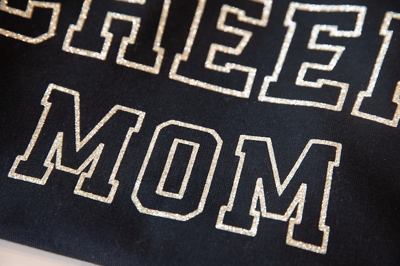 May include: A black shirt with the words "Cheer Mom" in silver glitter lettering.