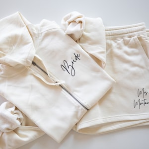 Puede incluir: Una sudadera con capucha de color crema con la palabra "Bride" bordada en la parte delantera y un par de pantalones cortos a juego de color crema con el texto "Mrs. Montano" bordado en la parte posterior.