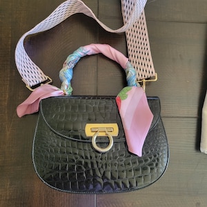 May include: Black, glossy handbag with a crocodile skin texture. The bag features a gold-colored clasp and a handle wrapped in a pink scarf. A patterned strap with black and white accents is attached.