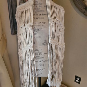 Puede incluir: Un chaleco blanco de crochet con flecos. El chaleco se muestra en un maniquí.