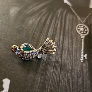 May include: A gold-tone bird brooch with a green gemstone and a detailed filigree design. The bird has a long tail with a fan-like shape.