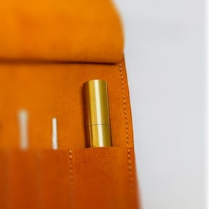 May include: A brown leather roll-up case with multiple slots for holding pens and pencils. A gold-colored pen is visible in one of the slots.