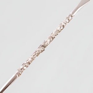 Op de afbeelding: Een zilveren metalen roerlepeltje met een decoratief swirl-patroon. De lepel heeft een afgerond uiteinde en een lange, dunne steel.