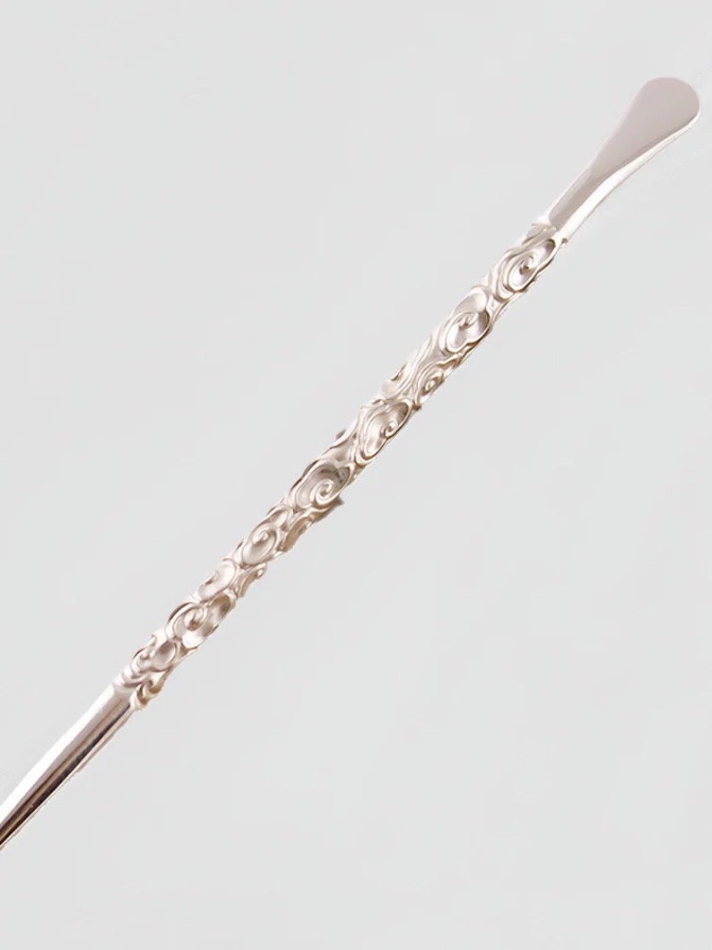 Op de afbeelding: Een zilveren metalen roerlepeltje met een decoratief swirl-patroon. De lepel heeft een afgerond uiteinde en een lange, dunne steel.