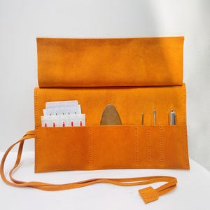 Könnte beinhalten: Eine offene, orangefarbene Wildleder-Rolltasche mit mehreren Taschen. Die Tasche enthält kleine Werkzeuge und Päckchen. Ein passender orangefarbener Krawattenverschluss sichert die Tasche. Die Tasche befindet sich auf einer weißen Oberfläche.