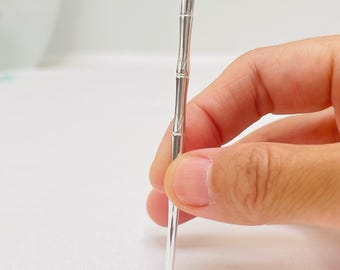 May include: A slender, silver-colored bamboo-shaped toothpick. The toothpick has a detailed design mimicking bamboo segments. The tip is pointed, and the overall design is sleek and modern. The toothpick is held by a hand against a white background.