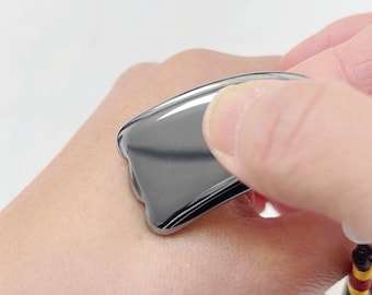 May include: A dark gray gua sha tool with a curved, smooth surface. The tool is held against the skin, suggesting its use for facial massage or skincare routines. The tool is made of a shiny, reflective material.
