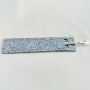 Op de afbeelding: Een grijze gevoerde etui met een zilveren, decoratief metalen gereedschap erin.