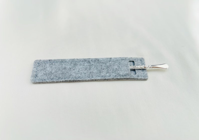Op de afbeelding: Een grijze gevoerde etui met een zilveren, decoratief metalen gereedschap erin.