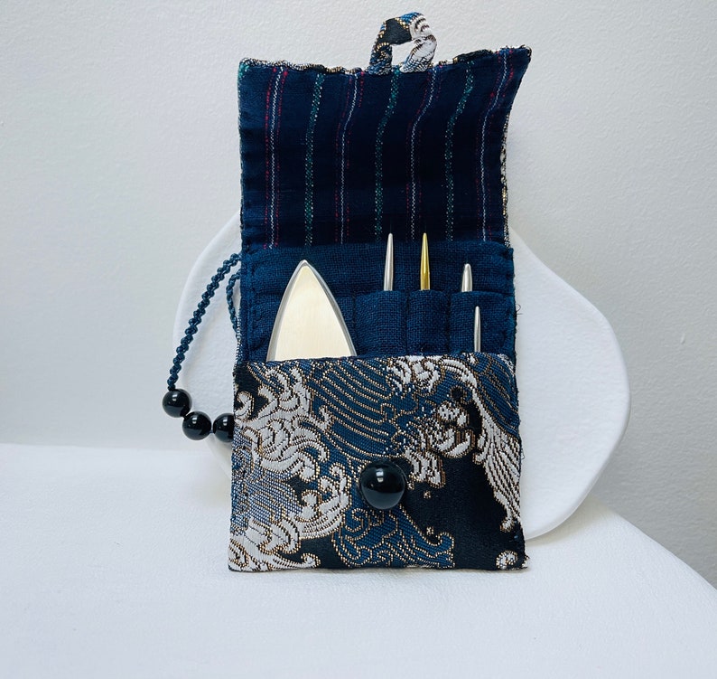 Op de afbeelding: Een klein, blauw en wit stoffen tasje met een ritssluiting. Het tasje is versierd met een golfpatroon en heeft een zwarte knoop. Het tasje is open en bevat een klein, zilveren gereedschap en drie naalden.