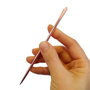 May include: A rose gold metal tool with a pointed tip and a rounded end. The tool is held in a hand.