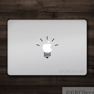 May include: A white Apple logo inside a lightbulb shape with a black outline. The lightbulb has a black spring at the bottom and black rays emanating from the top. The image is on a silver laptop with the text "©GRCDecals" in the bottom right corner.