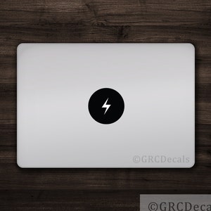 May include: A silver laptop with a black circular sticker displaying a white lightning bolt. The laptop rests on a dark wooden surface. The sticker is centrally placed. The bottom right corner of the image has the text "©GRC Decals".