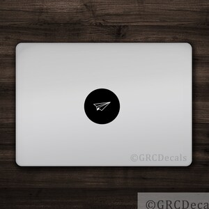Könnte beinhalten: Ein schwarzer runder Aufkleber mit einem weißen Papierflieger-Symbol auf einem silbernen Laptop.