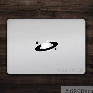 Puede incluir: Una pegatina en blanco y negro de una manzana con un anillo alrededor, que representa una galaxia, en un portátil plateado. La pegatina es un diseño simple con una manzana blanca y un anillo negro. El texto "©GRCDecals" está en la esquina inferior derecha de la imagen.