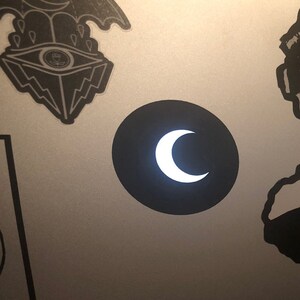 May include: A black circle sticker with a white crescent moon in the centre. The sticker is on a silver surface.