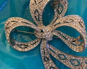 May include: A silver-toned brooch in the shape of a bow with rhinestones. The bow is made of multiple layers of ribbon with a detailed design.
