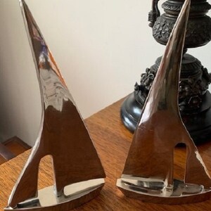 May include: Two silver sailboat sculptures on a wooden surface. The sailboats are made of metal and have a polished finish.