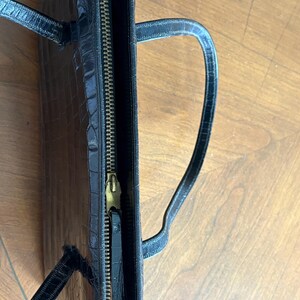May include: Black handbag with a crocodile skin texture. The bag has a gold zipper and two handles. The bag is open, revealing the interior. The bag is sitting on a wooden surface.