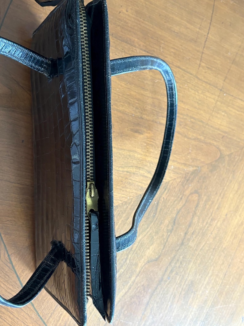 May include: Black handbag with a crocodile skin texture. The bag has a gold zipper and two handles. The bag is open, revealing the interior. The bag is sitting on a wooden surface.