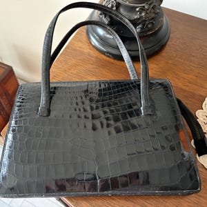 May include: Black rectangular handbag with two top handles. The bag has a textured, crocodile-like pattern. The handles are thin and black. The bag is sitting on a wooden surface.