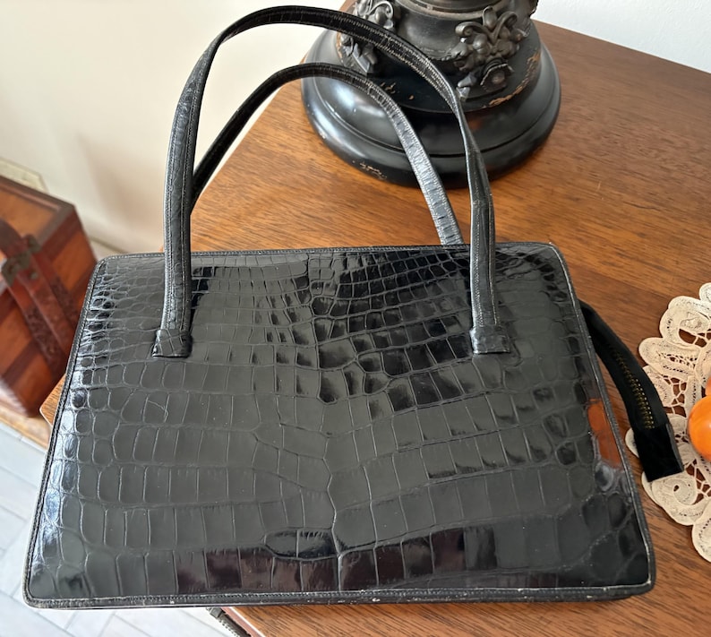 May include: Black rectangular handbag with two top handles. The bag has a textured, crocodile-like pattern. The handles are thin and black. The bag is sitting on a wooden surface.