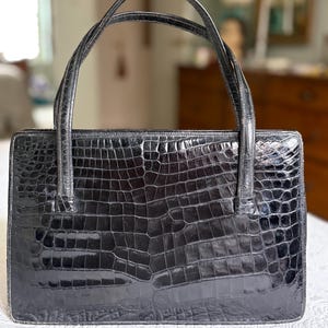 May include: A black, rectangular handbag with a crocodile skin texture. The bag has two curved handles and a structured shape. The glossy finish reflects light, highlighting the detailed pattern. The bag is a classic accessory.