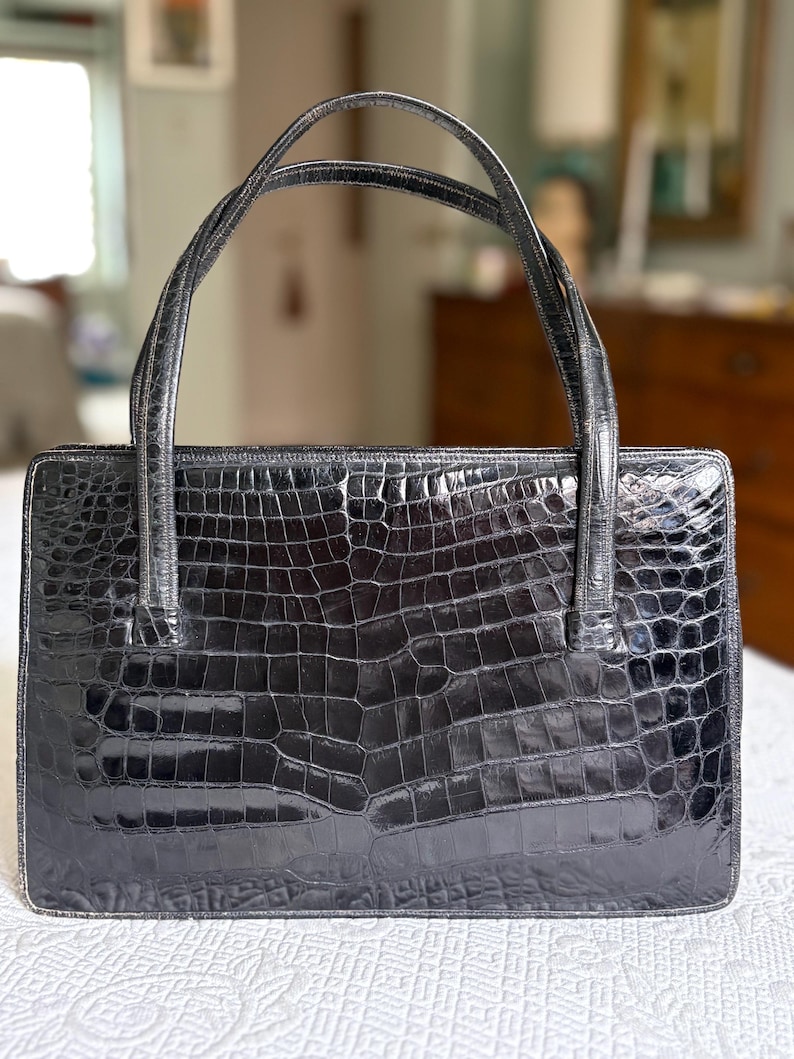 May include: A black, rectangular handbag with a crocodile skin texture. The bag has two curved handles and a structured shape. The glossy finish reflects light, highlighting the detailed pattern. The bag is a classic accessory.